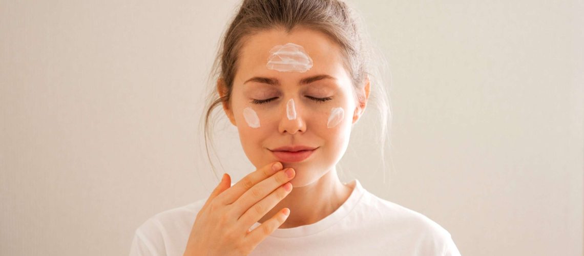 Young woman with dabs of cream on her forehead, cheeks, and nose, applying moisturizer with her eyes closed.
